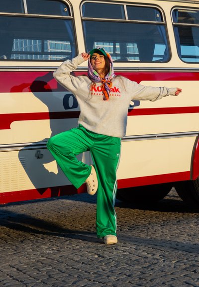 Sudadera gris con el logo de "Kodak", pantalones verdes de pierna ancha, bufanda colorida con estampado y zuecos crema, posando junto a un autobús vintage.