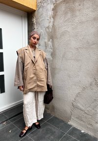 Gilet beige oversize con dettagli nei bottoni sopra una maglietta grigia a maniche lunghe, pantaloni bianchi a gamba larga e scarpe marroni scure, mentre tiene una borsa scura.