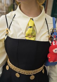 Corbata amarilla con estampado, con un pin dorado sobre una camisa crema, vestido negro con tirantes finos y cinturón de monedas doradas, sosteniendo un bolso azul con un llavero de peluche rojo.