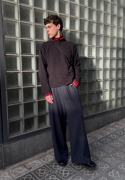 Joven con jersey oscuro de rayas y pantalones anchos negros, de pie con la mano en el bolsillo, mirando hacia un lado, frente a una pared de bloques de vidrio.