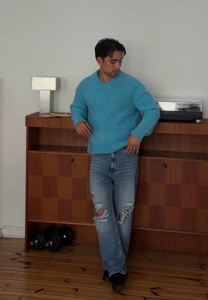 Homme en pull bleu et jean déchiré, debout près d'une armoire en bois avec des haltères et une platine dessus.