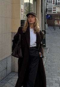 Femme portant un manteau foncé, une chemise blanche, un pantalon à rayures fines et une casquette noire, debout sur un trottoir en ville tenant un grand sac à main foncé.