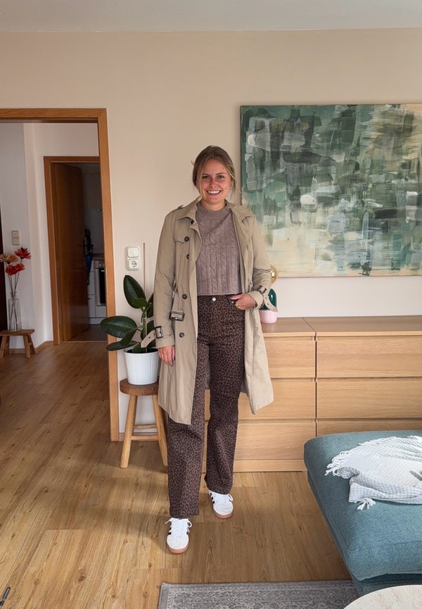 Joven mujer con abrigo trench beige, pantalones estampados y zapatillas blancas, de pie en un salón moderno con plantas y arte mural abstracto.