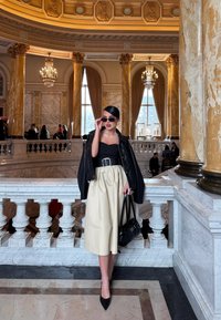 Μαύρο δερμάτινο μπουφάν, μαύρο τοπ χωρίς straps, κρεμ σατέν μίντι φούστα, μυτερά μαύρα τακούνια, μαύρη τσάντα, με γυαλιά ηλίου, σε έναν μεγαλοπρεπή εσωτερικό χώρο.