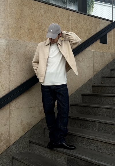 Chaqueta texturizada beige claro sobre una camiseta de cuello redondo blanca, combinada con jeans azul oscuro y botas negras. Una gorra gris completa el look.