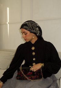 Blouse noire côtelée avec des boutons en or et perle, associée à une pochette noire à motif floral avec une chaîne dorée et une jupe grise.