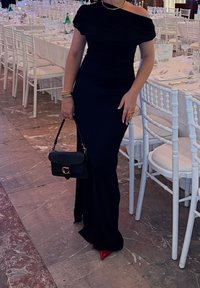 Femme en robe noire à épaules dénudées tenant un sac à main noir, debout à côté d'une table à manger dressée avec des chaises blanches dans une pièce au sol en marbre.