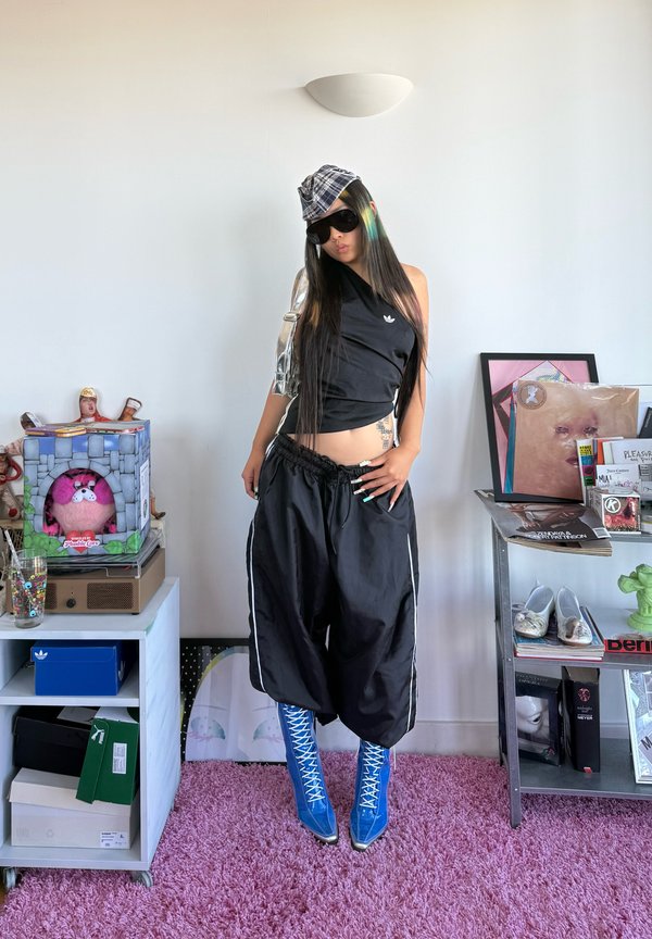 Jeune personne portant un haut noir asymétrique, un pantalon noir ample, des bottes bleues à lacets, un chapeau à carreaux et des lunettes de soleil, se tient sur un tapis violet dans une pièce avec des étagères.