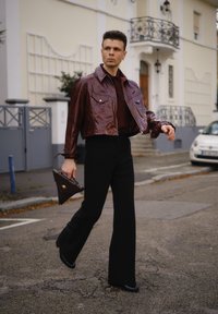 Muž kráča po ulici v lesklom gaštanovom skrátenom saku, čiernych rozšírených nohaviciach a drží trojuholníkovú tmavohnedú kabelku.