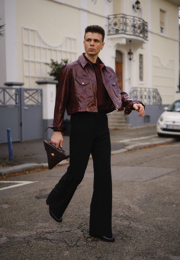 Mann, der auf der Straße geht, trägt eine glänzende kastanienbraune, verkürzte Jacke, schwarze, ausgestellte Hosen und hält eine dreieckige, dunkelbraune Handtasche.