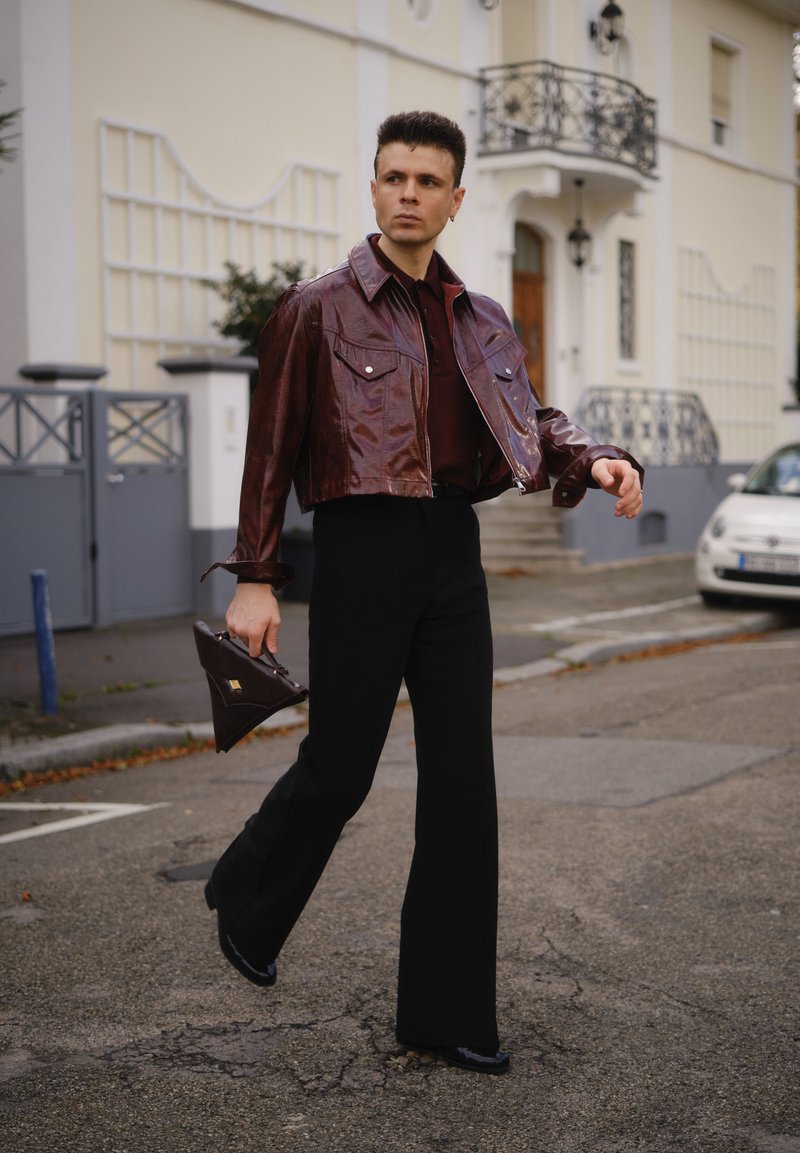 Muž kráča po ulici v lesklom gaštanovom skrátenom saku, čiernych rozšírených nohaviciach a drží trojuholníkovú tmavohnedú kabelku.