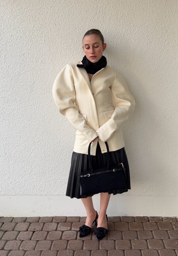 Femme portant une veste crème à manches bouffantes, jupe plissée noire, foulard noir, tenant un sac à main noir, debout contre un mur blanc texturé.