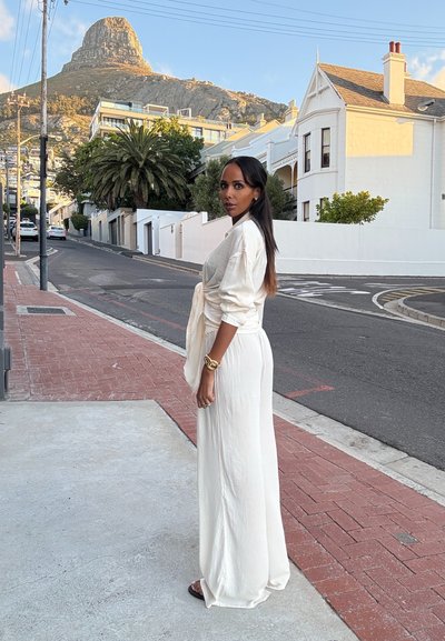 Largo vestido blanco fluido con un diseño envolvente y mangas casuales. La modelo está de pie en una acera con un fondo de palmeras y una montaña.