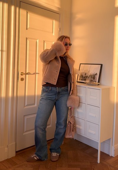 Chaqueta de piel sintética beige con mangas largas, top marrón y vaqueros acampanados azul claro. Los accesorios incluyen bolso rosa y gafas de sol redondas.