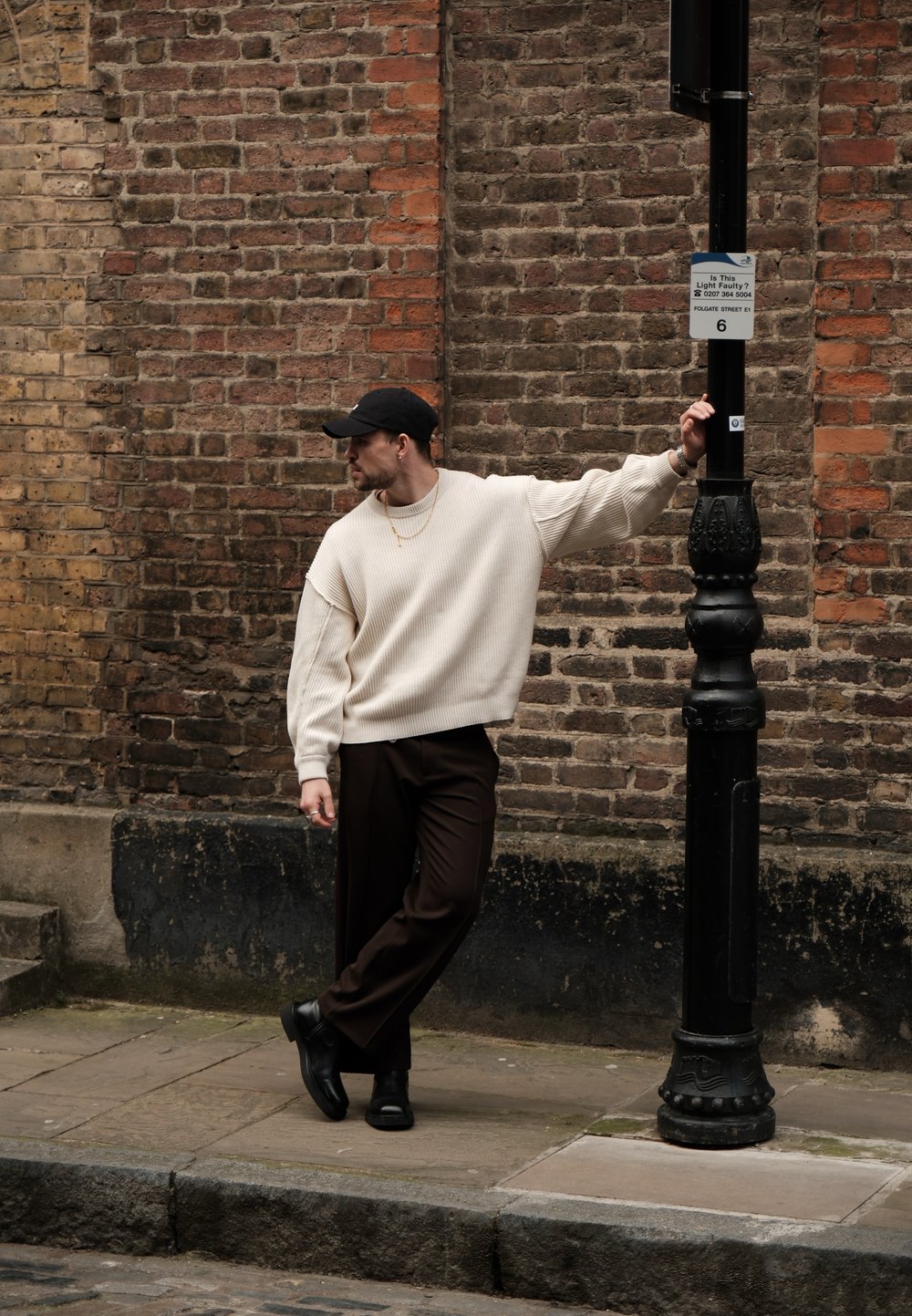 Pulover crem cu striații, pantaloni maro și pantofi negri. Stă lângă un stâlp de iluminat negru, sprijinindu-se casual de un zid de cărămidă.