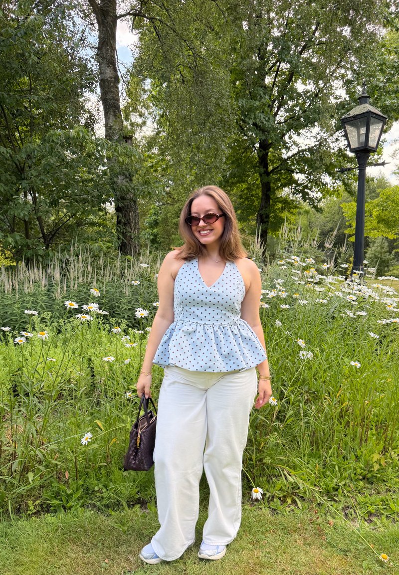 Jeune femme portant des lunettes de soleil, un haut bleu à pois et un pantalon blanc, souriante dans un jardin avec des marguerites et des arbres.