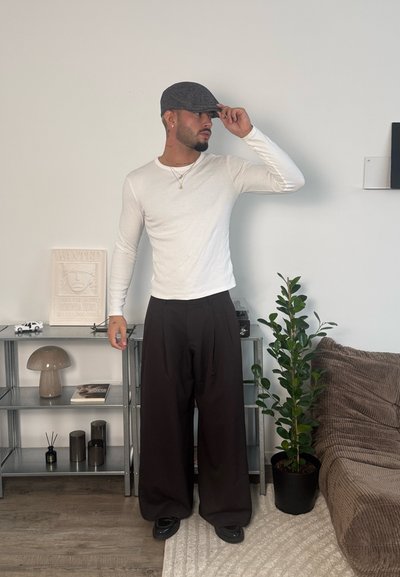 Hombre con camisa blanca de manga larga, pantalones negros amplios y gorra plana, de pie en el interior junto a una estantería con objetos decorativos y una planta en maceta al lado de un sofá marrón.