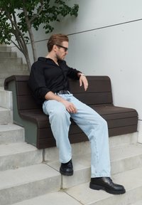 Camisa negra de botones, jeans azul claro y zapatos negros. Sentado en un banco de madera oscura contra una pared clara y vegetación.