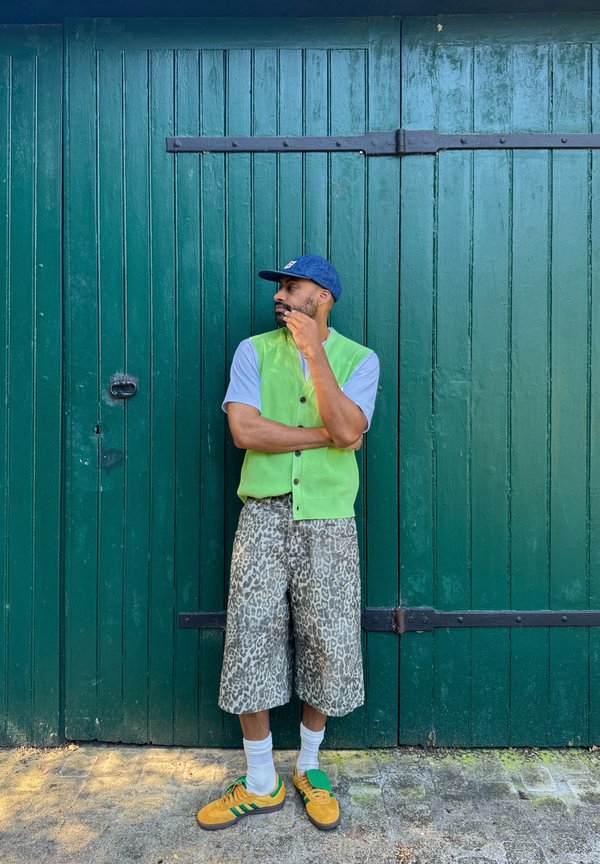 Man wearing a green vest, grey T-shirt, and leopard print shorts, standing in front of a green wooden door with black fittings.
