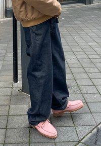 Personne portant un pantalon ample bleu foncé et des chaussures en cuir roses, debout sur un trottoir pavé gris, tenant des clés près d'un poteau.