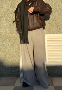 Giacca di pelle marrone, pantaloni della tuta grigi con strisce bianche ai lati, sciarpa nera e scarpe nere, in piedi contro un muro texturizzato.