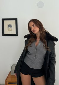 Schwarzer Ledermantel mit Fellkragen, grauer Cardigan und schwarze Shorts. Glatte Textur und figurbetontes Design, vor einem minimalistischen Hintergrund.