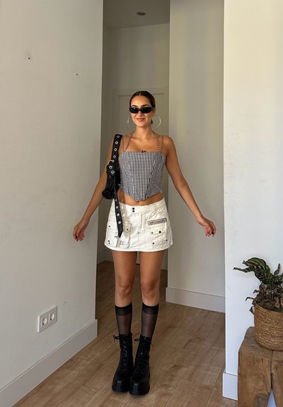 Mujer vistiendo una blusa a cuadros blancos y negros, una mini falda blanca, botas negras, medias transparentes, gafas de sol y grandes pendientes de aro, de pie en una habitación.