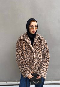 Καφέ παλτό από τεχνητή γούνα με λεοπάρ σχέδιο, φαρδύ γιακά, μακριά μανίκια και μαύρα κουμπιά, συνδυασμένο με μια μαύρη τσάντα και ανοιχτόχρωμα γυαλιά ηλίου.