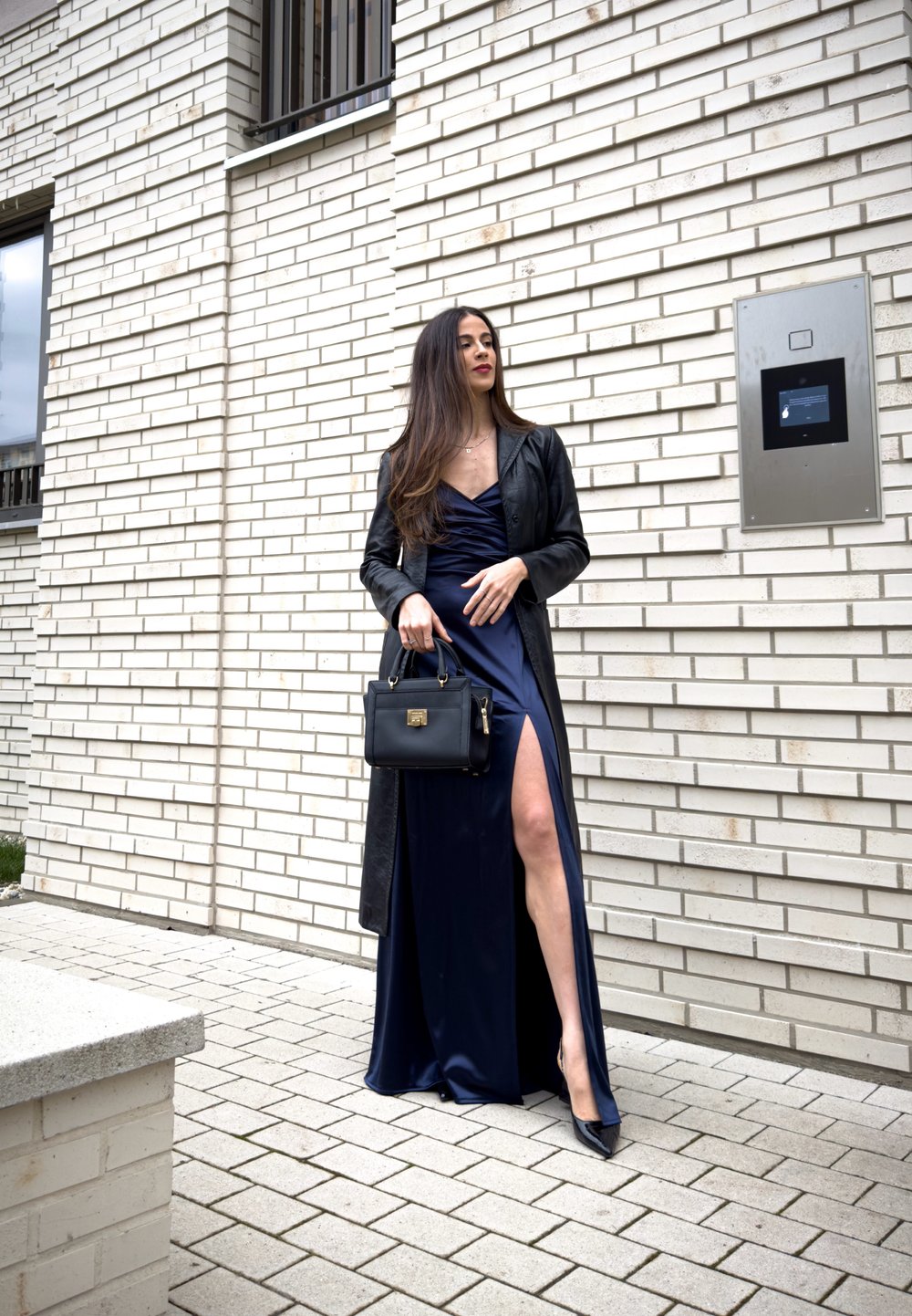 Femme en robe bleu marine fendue jusqu'à la cuisse, manteau en cuir noir et talons, tenant un sac à main noir, debout devant un mur de briques blanches.