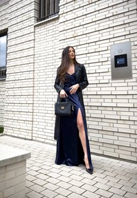 Femme en robe bleu marine fendue jusqu'à la cuisse, manteau en cuir noir et talons, tenant un sac à main noir, debout devant un mur de briques blanches.