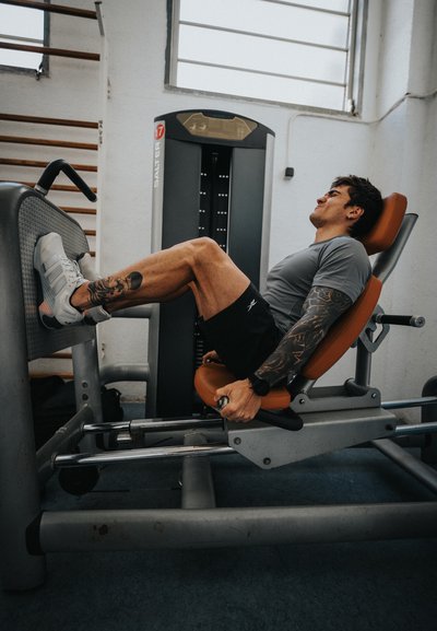 Hombre con tatuajes empujando máquina de prensa de piernas con peso en un gimnasio luminoso con una gran ventana y equipo de fitness.