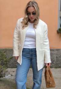 Femme portant des lunettes de soleil, une veste blanche texturée, une chemise blanche, un jean bleu, tenant un sac à main en daim marron, debout contre un mur pêche.