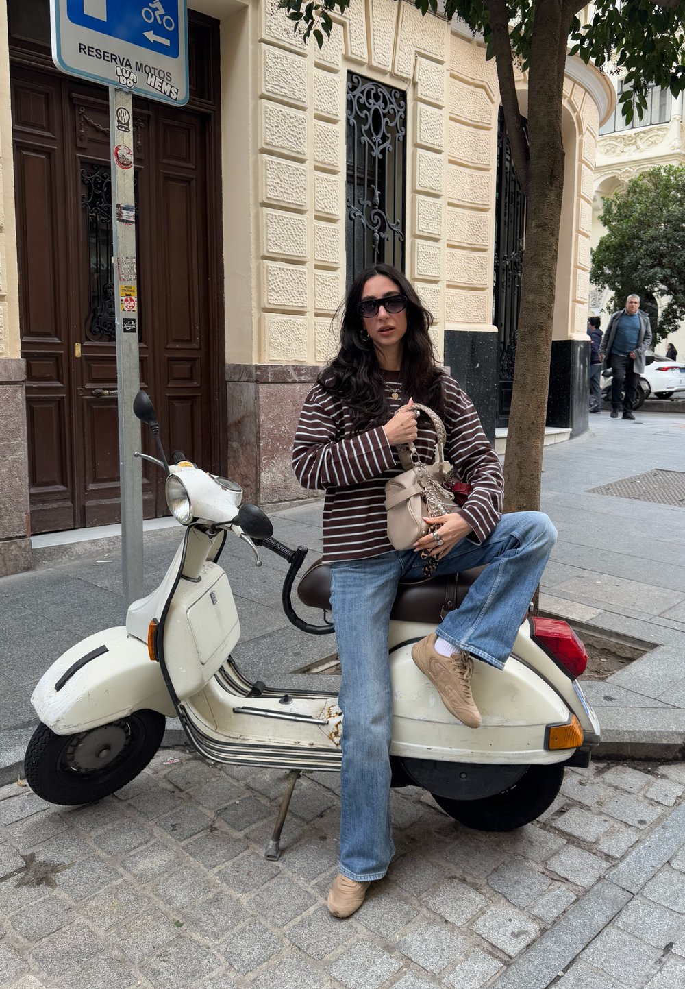 Scooter bianco con un design vintage su una strada lastricata. Camicia a righe marroni e bianche, jeans blu e sneakers beige. Porta marrone sullo sfondo.