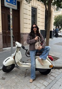 Scooter bianco con un design vintage su una strada lastricata. Camicia a righe marroni e bianche, jeans blu e sneakers beige. Porta marrone sullo sfondo.