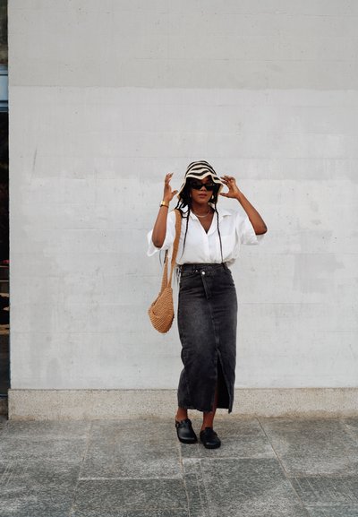 Camisa blanca de gran tamaño, falda midi de mezclilla negra, bolso de mimbre color marrón, zuecos negros y un sombrero de cubo a rayas. Superficies suaves y líneas definidas.