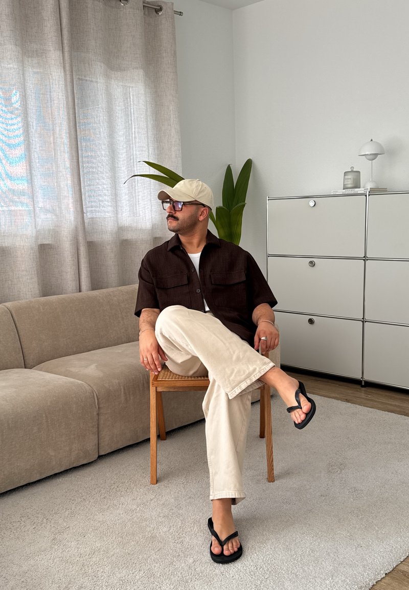 Una persona è seduta su una sedia di legno, indossando una camicia a maniche corte marrone scuro, pantaloni beige, ciabatte nere e un cappellino beige chiaro in una stanza luminosa.