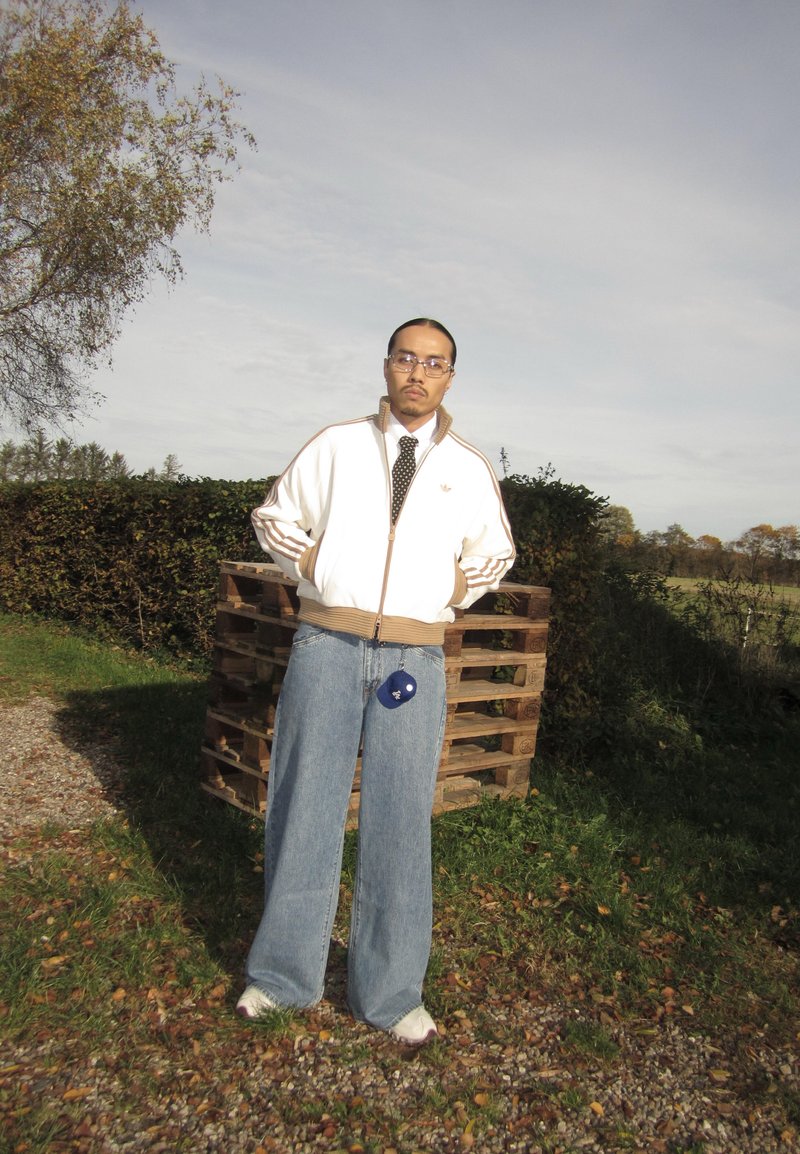 Giacca bianca con zip e dettagli marroni, abbinata a jeans blu oversize. Accessori includono una borsetta blu. Ambiente naturale all'aperto.