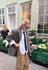 Pārāk liels brūns žakets virs baltas krekla, melna kaklasaites un gaišbrūnas platās bikses; nēsā saulesbrilles, tur melnu somu pie tirgus.