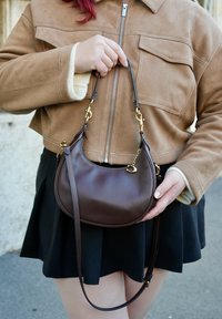 Ruda odinė peties rankinė su išlenkta forma, auksinėmis detalių ir nuimamu dirželiu, laikoma rankoje priešais rudą zomšinį švarką.