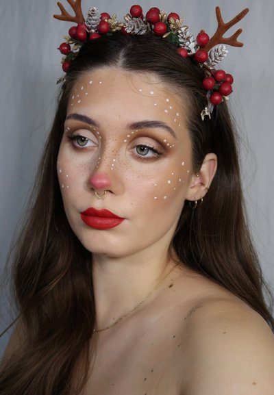 Modelo con una diadema floral con bayas rojas y cuernos, con nariz roja, pecas blancas y un llamativo lápiz labial rojo sobre un fondo neutro.