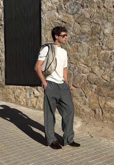 Camiseta de manga corta gris claro con textura acanalada, pantalones holgados gris oscuro, zapatos oscuros y un bolso de hombro beige con ribete negro, de pie contra una pared de piedra.