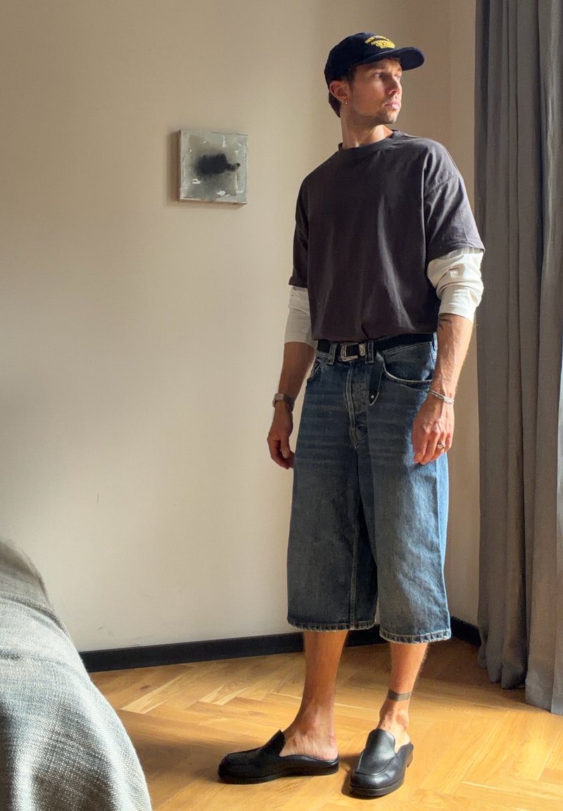 Homme portant une casquette noire, des chemises superposées, un long short en jean et des chaussures noires slip-on, debout dans une pièce baignée de soleil près d'un rideau et d'une œuvre murale abstraite.