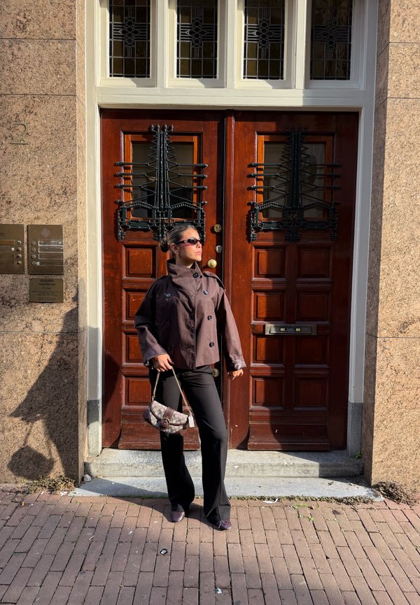 Woman in sunglasses and brown jacket stands on brick sidewalk in front of large wooden double doors with decorative black metalwork.