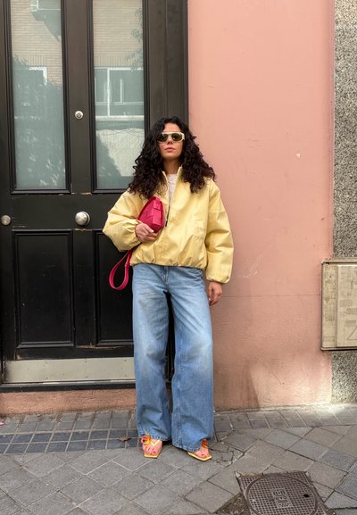 Mujer con cabello rizado que lleva una chaqueta amarilla, vaqueros de pierna ancha, sandalias rosas y gafas de sol, sosteniendo un bolso rojo, de pie junto a una puerta negra y una pared rosa.