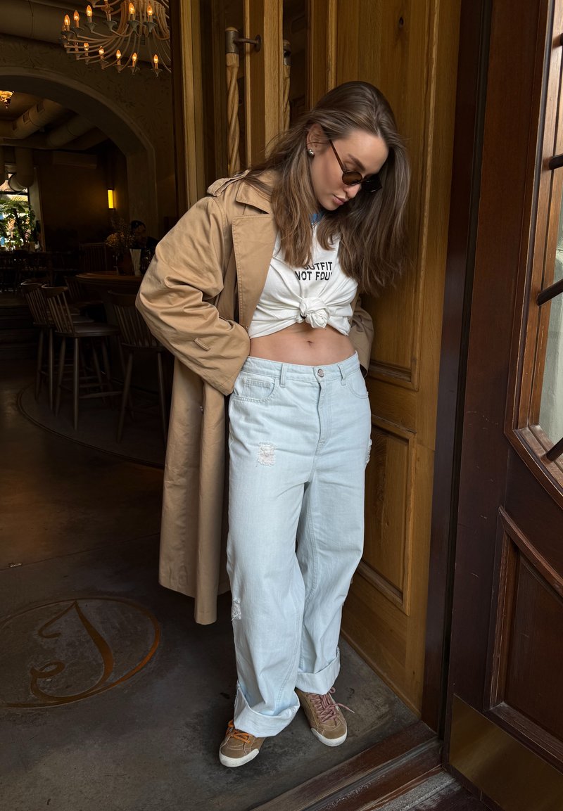 Femme avec des lunettes de soleil portant un trench-coat beige, un crop top blanc noué, un jean bleu clair ample et des baskets marron, debout près d'une porte en bois.