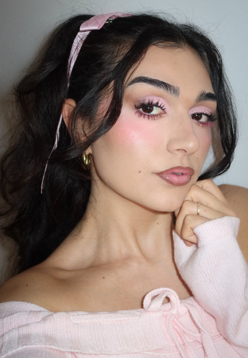 Jeune femme aux cheveux foncés attachés par un ruban rose, portant un fard à paupières rose et un pull, posant avec la main près du menton sur un fond uni.
