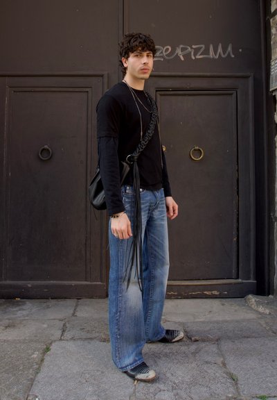 Camisa negra de manga larga, vaqueros de pierna ancha en color azul claro y bolso negro con detalles de flecos. Los zapatos tienen un patrón a rayas. Fondo urbano.