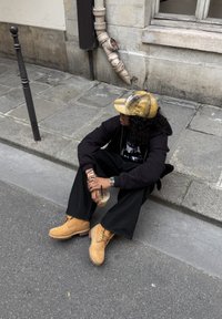 Persona dzeltenā rūtainā cepurē, melnā kapučjakā, melnās biksēs un brūnās zābakos, sēžot pie apmalītes, turot ledus kafiju pilsētas ielā.