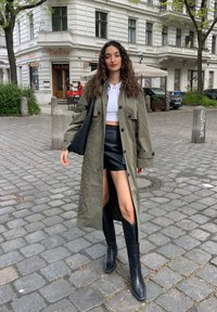 Cappotto lungo verde oliva con grandi bottoni, abbinato a un crop top bianco e una minigonna di pelle nera. Stivali neri fino al ginocchio completano il look.