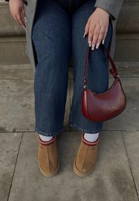 Botas de tornozelo em camurça castanha com meias realçadas em vermelho, jeans retos azuis e uma pequena bolsa de couro burgundy. Fundo de pedra texturizada.